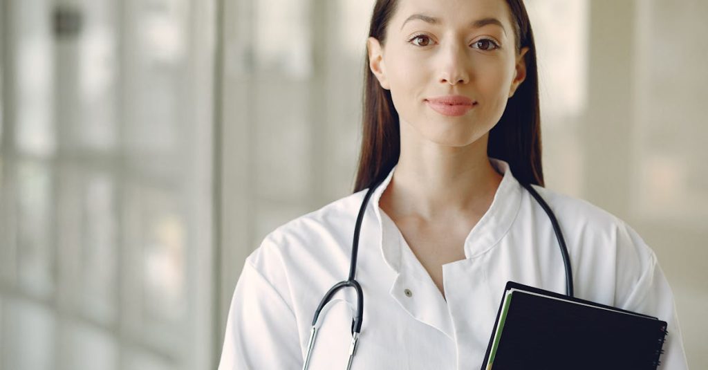 Crop smiling young ethnic female doctor in medical uniform with stethoscope and notebook standing in modern medical room with panoramic window